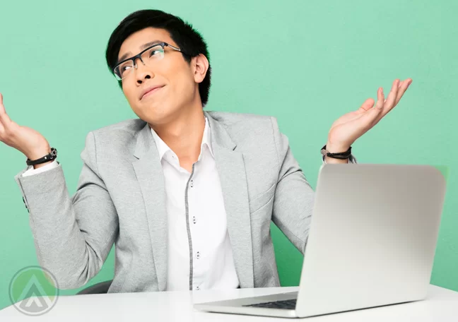 asian-male-employee-in-glasses-shrugging-in-front-of-laptop-computer