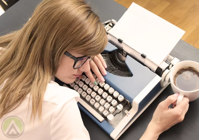 female-writer-in-glasses-resting-on-typewriter-holding-coffee-mug