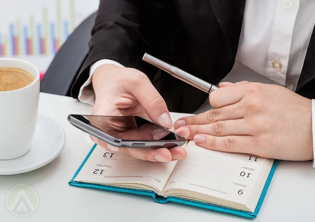 businessman-hands-using-smartphone-writing-on-calendar-planner-notepad-with-coffee